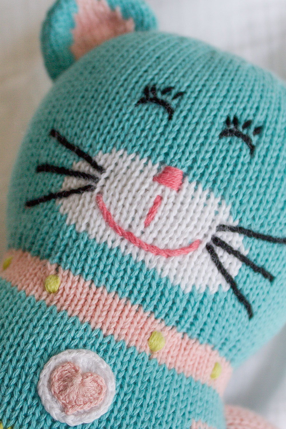 A close-up of a knitted toy cat with turquoise, white, and pink yarn, featuring stitched facial features and a heart detail on its chest.