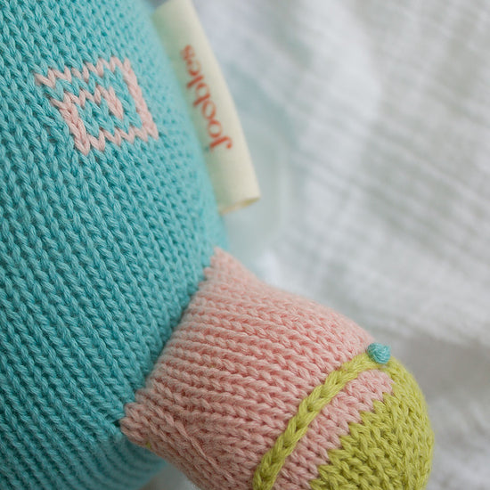 A close-up of a knitted stuffed toy with a blue body, pink and green limb, and a small label reading joolz. The toy is lying on a textured white fabric.