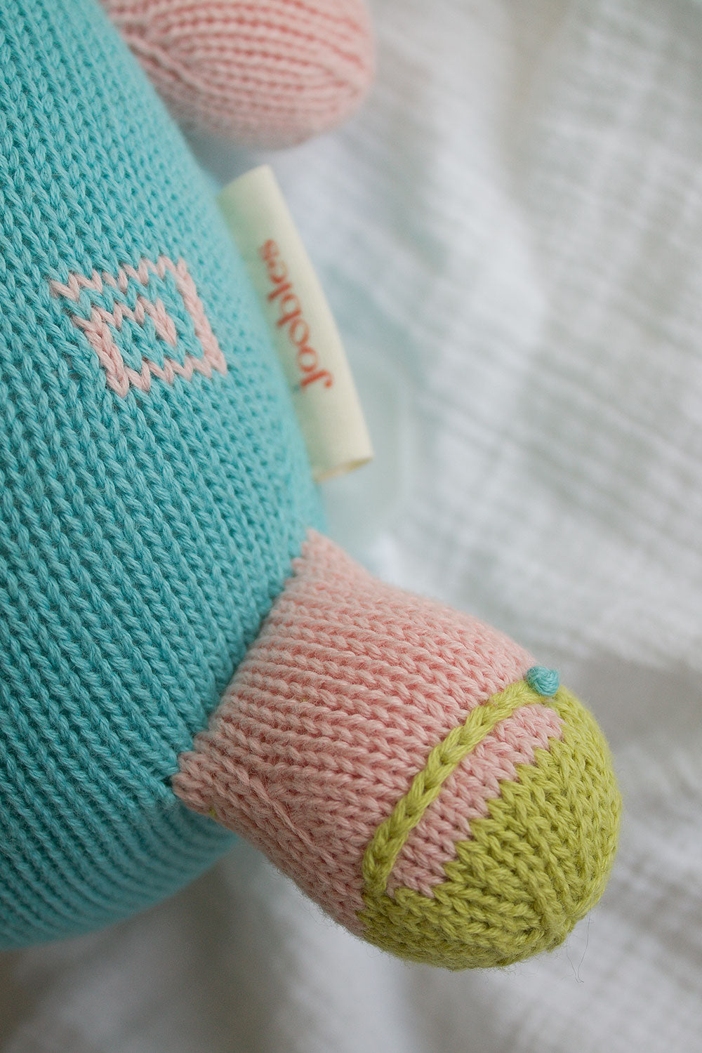 A close-up of a knitted stuffed toy with a blue body, pink and green limb, and a small label reading joolz. The toy is lying on a textured white fabric.
