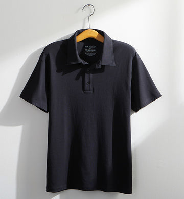 A black short-sleeve polo shirt hangs on a wooden hanger against a white wall, illuminated by natural light.