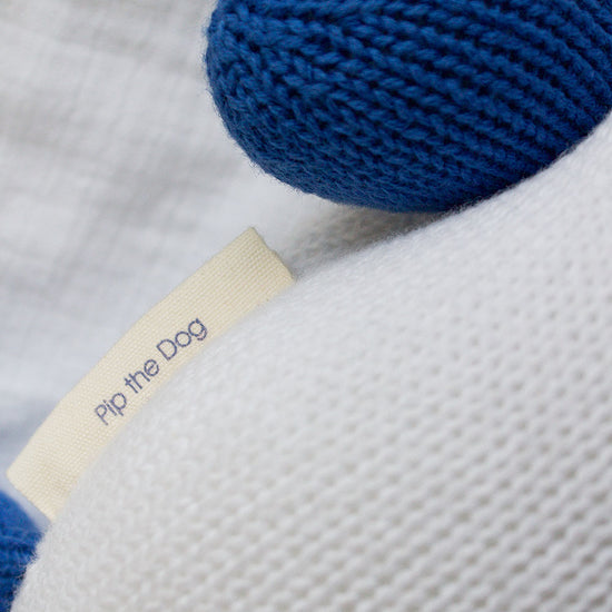 A close-up of a knitted stuffed animal with blue ears and a fabric tag that reads Pip the Dog.