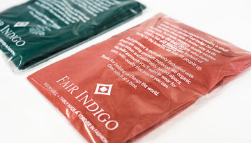 Two Fair Indigo t-shirts, one red and one green, are packaged in clear plastic bags with sustainability messaging and branding printed on the front.