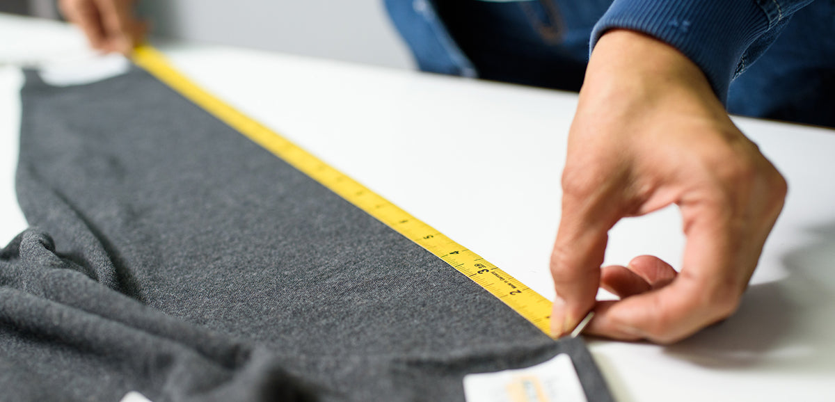 Person measuring a dark grey fabric garment with a yellow measuring tape on a white surface.