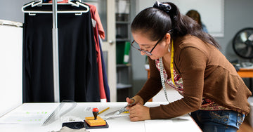 A woman with glasses and a measuring tape around her neck uses scissors to cut fabric on a table in a workspace with clothing and documents nearby.
