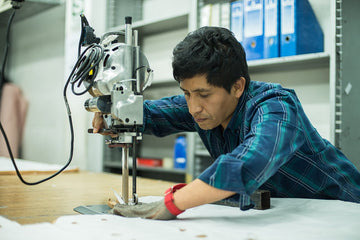 A man uses an industrial fabric cutting machine to cut white cloth on a worktable in a workshop with shelves and folders in the background.