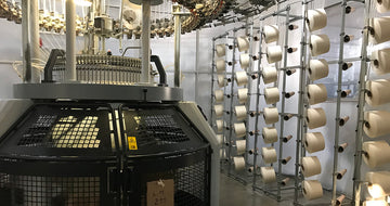 Large industrial knitting machine with multiple spools of white yarn arranged on racks in a factory setting.