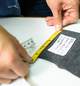 A person measures a piece of dark fabric with a yellow measuring tape; a white label with handwritten notes is on the fabric.