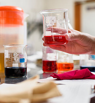 A person holds a beaker containing red liquid, with other beakers filled with colored liquids on a lab table.